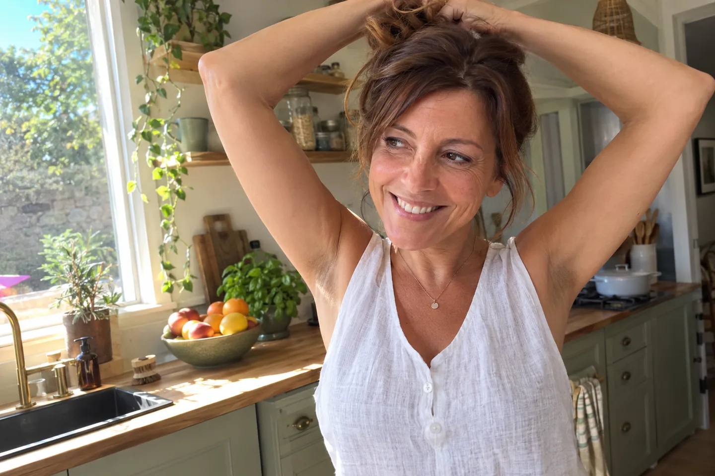 Femme en débardeur qui attache ses cheveux dans une cuisine ensoleillée, bras levés