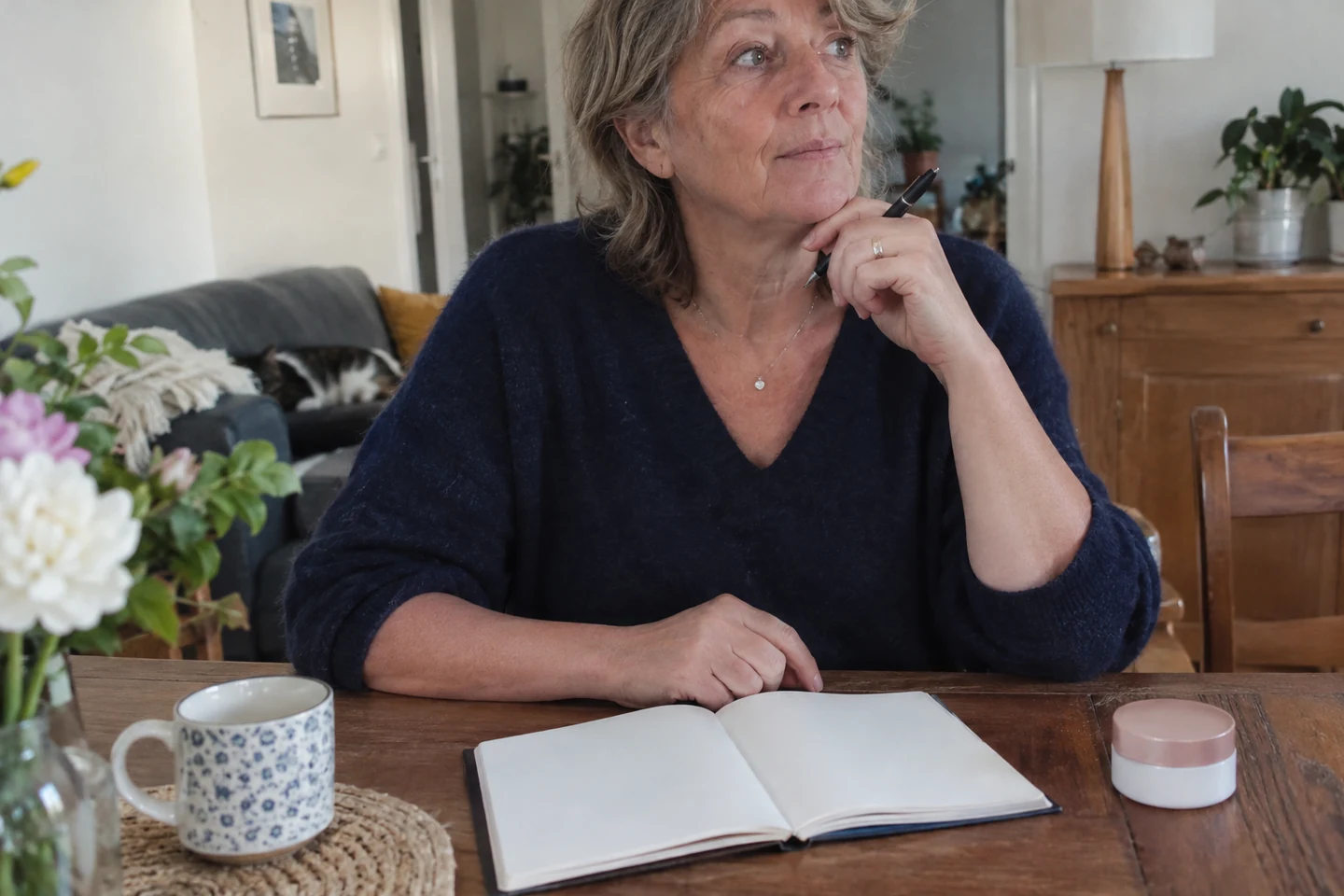 Femme assise devant un carnet ouvert vierge, tenant un stylo près d’un pot fermé.