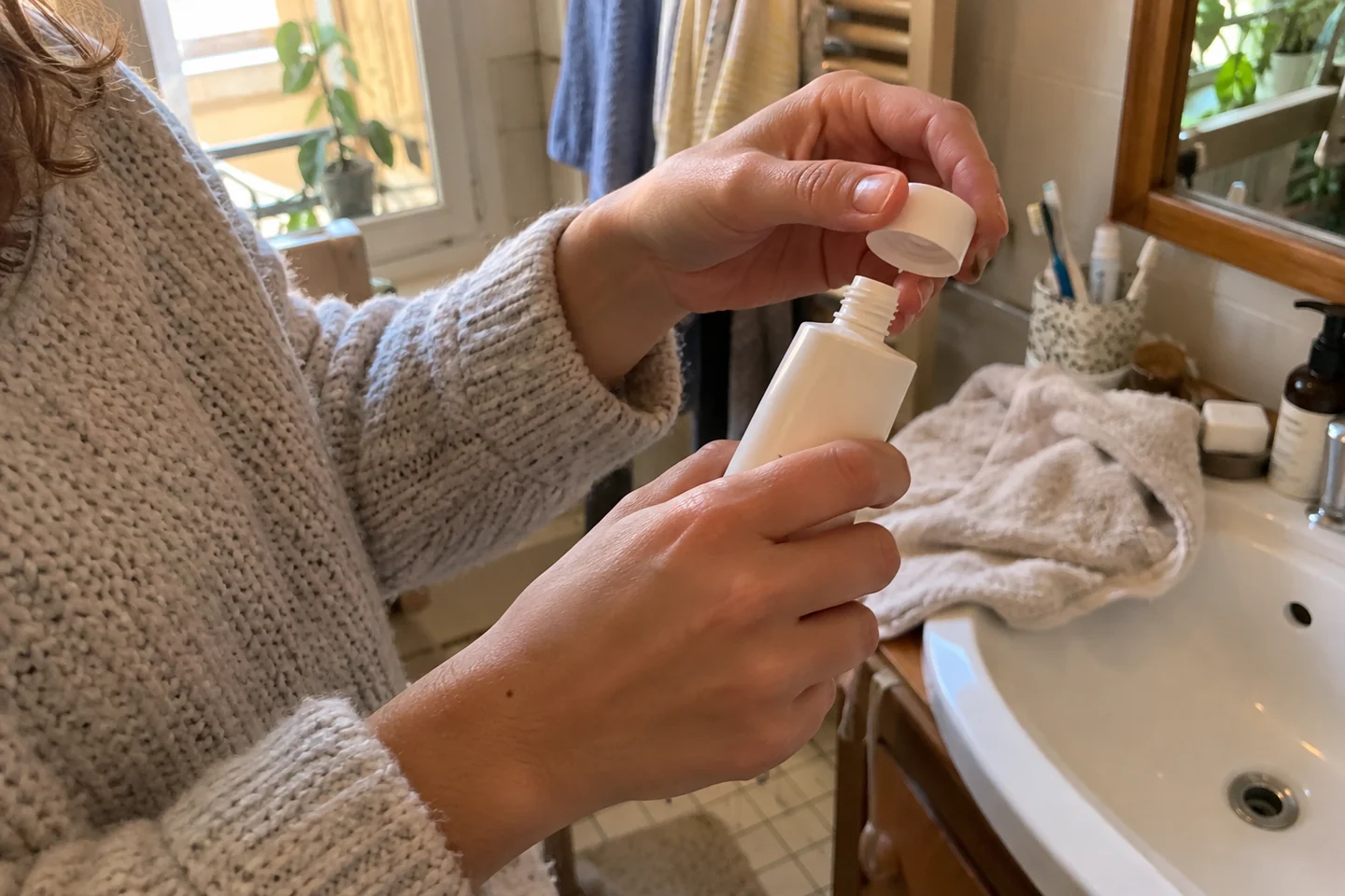 Deux mains retirent le bouchon d’un contenant de soin près d’un lavabo.